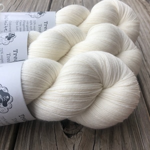 May include: Close-up of two skeins of white yarn. The yarn is tightly wound and has a soft, textured appearance. The label reads "Trea" and "White". The yarn is likely used for knitting or crocheting projects.