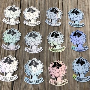 May include: A collection of pirate sheep stickers. Each sticker features a fluffy white sheep wearing a black pirate hat with a skull and crossbones. The word "YARRRRRN" is printed below each sheep in a banner. The stickers have a holographic sheen.