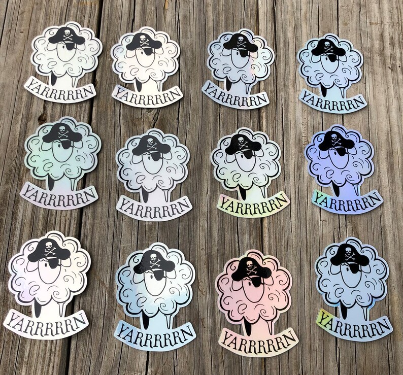 May include: A collection of pirate sheep stickers. Each sticker features a fluffy white sheep wearing a black pirate hat with a skull and crossbones. The word "YARRRRRN" is printed below each sheep in a banner. The stickers have a holographic sheen.