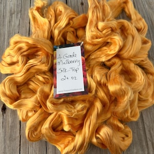 May include: A pile of golden-yellow A-Grade Mulberry Silk Top fiber. A small tag reads "A-Grade Mulberry Silk Top 2+ oz" with a website address. The silk fiber is soft and appears ready for spinning or other fiber arts projects.