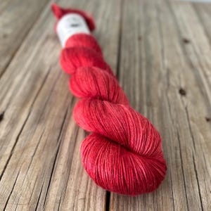 May include: A skein of vibrant red yarn, tightly wound and ready for crafting. The yarn is a rich, solid color, with a slight sheen, and is displayed on a rustic wooden surface. The label is white with text. Perfect for knitting or crochet projects.
