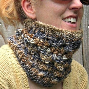May include: A hand-knitted cowl in shades of brown, blue, and cream. The chunky knit scarf is worn around the neck and is made of a soft, textured yarn. The cowl is a cosy accessory for cold weather.