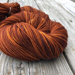May include: Close-up of a skein of yarn in shades of brown and orange. The yarn is tightly wound, with visible strands and color variations. The yarn is resting on a weathered wooden surface.