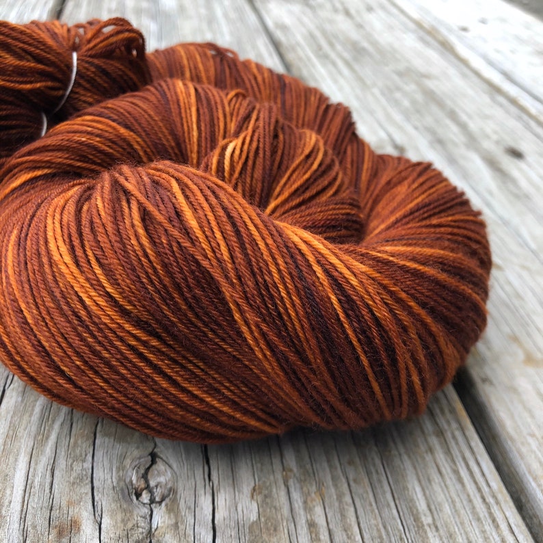 May include: Close-up of a skein of yarn in shades of brown and orange. The yarn is tightly wound, with visible strands and color variations. The yarn is resting on a weathered wooden surface.