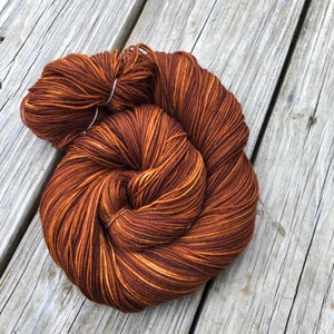 May include: A skein of brown yarn with streaks of lighter brown and gold. The yarn is tightly wound and secured with a white tag. The yarn is likely for knitting or crocheting projects.