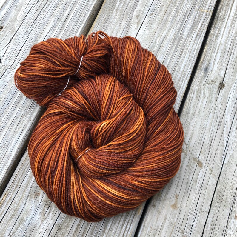 May include: A skein of brown yarn with streaks of lighter brown and gold. The yarn is tightly wound and secured with a white tag. The yarn is likely for knitting or crocheting projects.