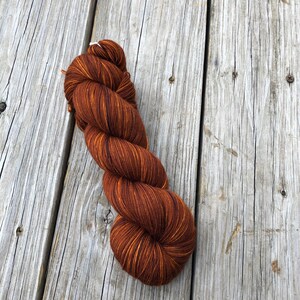 May include: A skein of brown yarn with golden highlights, resting on a weathered wooden surface. The yarn is tightly twisted, showcasing a gradient of warm, earthy tones. Ideal for knitting or crochet projects.