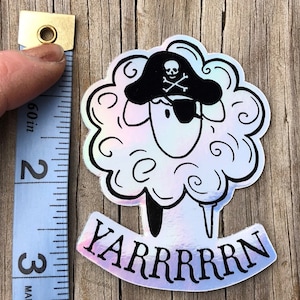 May include: A holographic sticker featuring a sheep wearing a pirate hat and eye patch. The sticker has the text "YARRRRRN" in black. The sticker is approximately 3 inches tall.