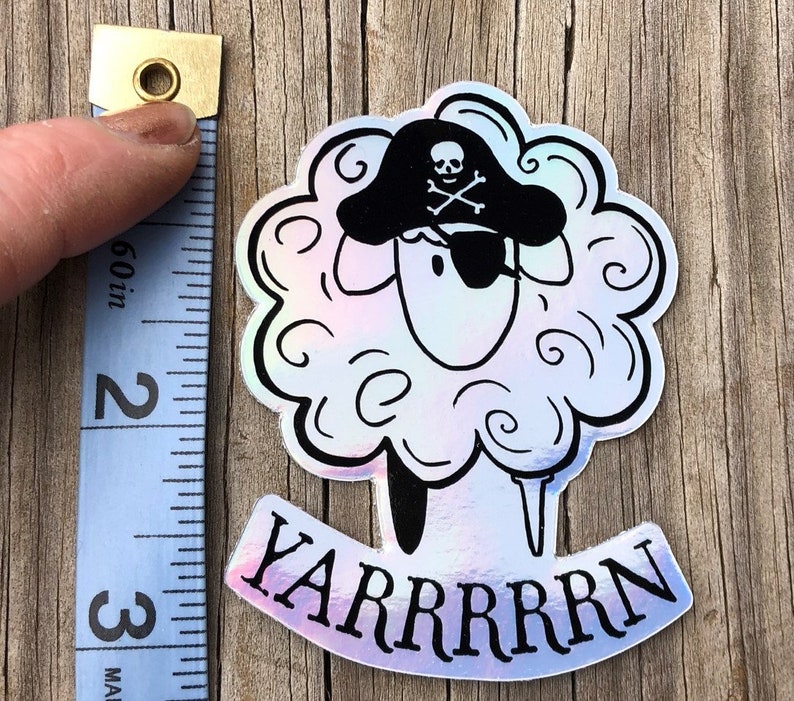 May include: A holographic sticker featuring a sheep wearing a pirate hat and eye patch. The sticker has the text "YARRRRRN" in black. The sticker is approximately 3 inches tall.