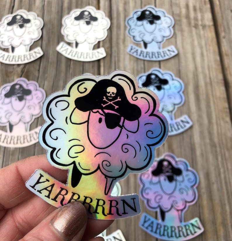 May include: Holographic stickers featuring a cartoon sheep wearing a pirate hat with a skull and crossbones. The sheep has fluffy, cloud-like wool and the word "YARRRRRN" is written below. The stickers have a rainbow sheen and are on a wooden surface.