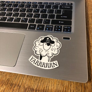 May include: A black and white sticker featuring a cartoon sheep wearing a pirate hat with a skull and crossbones. The sticker has the text "YARRRRRN" in a banner below the sheep. The sticker is on a laptop.