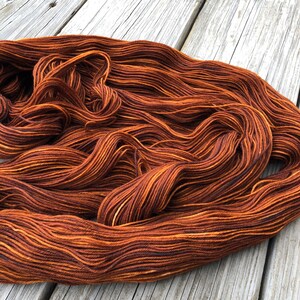 May include: A skein of hand-dyed yarn in shades of brown and orange, with subtle variations in color. The yarn is arranged in loose, flowing waves, showcasing its texture and color gradients. The yarn is likely for knitting or crocheting projects.