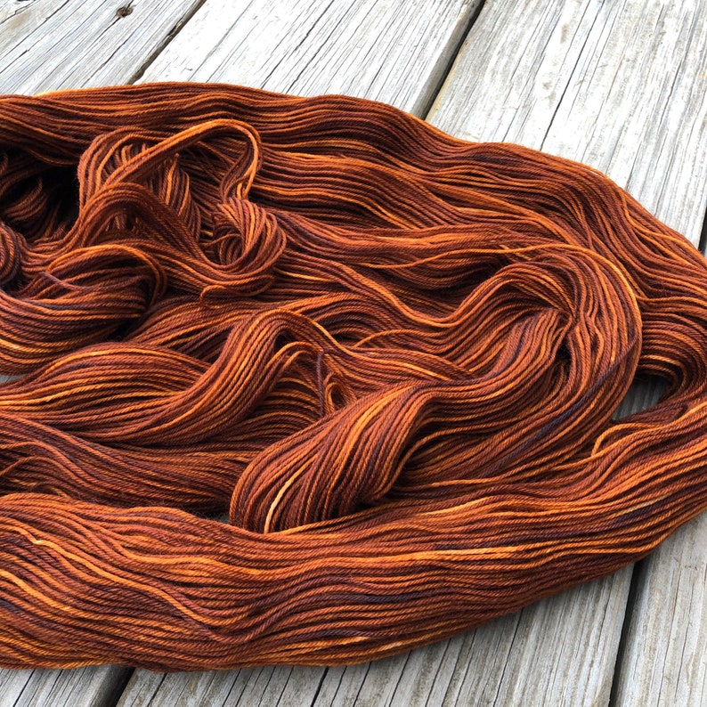May include: A skein of hand-dyed yarn in shades of brown and orange, with subtle variations in color. The yarn is arranged in loose, flowing waves, showcasing its texture and color gradients. The yarn is likely for knitting or crocheting projects.