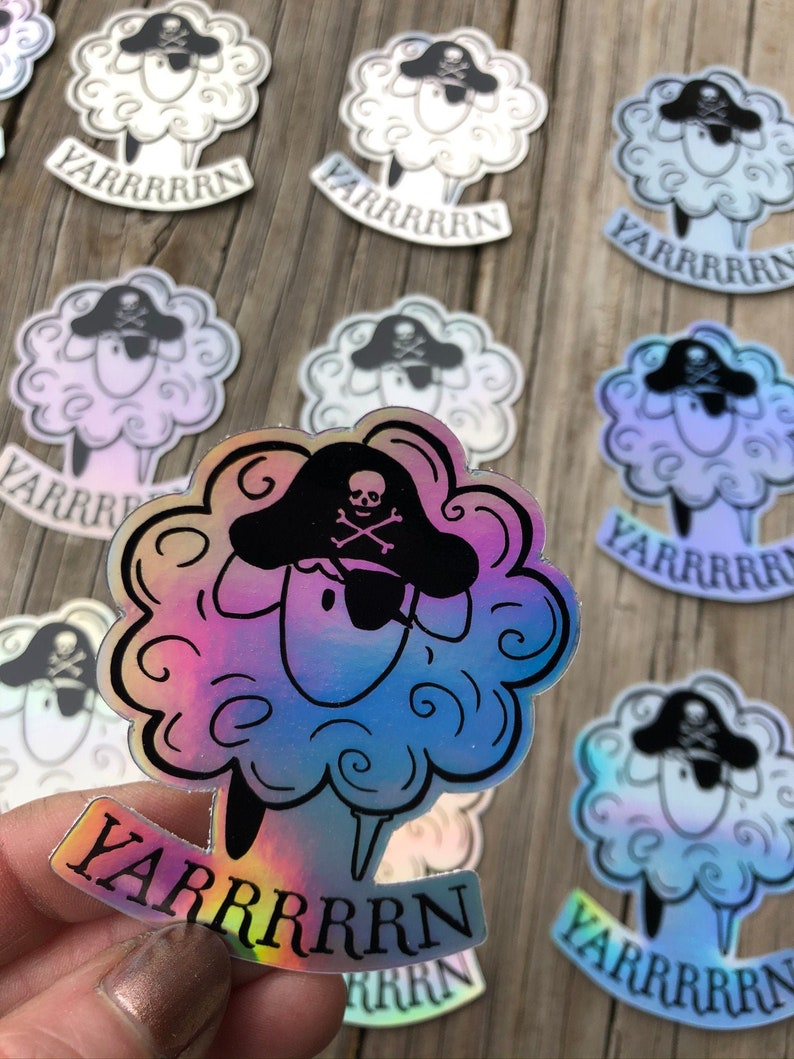 May include: A collection of stickers featuring a sheep with a pirate hat and the word "YARRRRRN". The sheep has a fluffy, cloud-like body and a black pirate hat with a skull and crossbones. The stickers have a holographic, rainbow sheen.