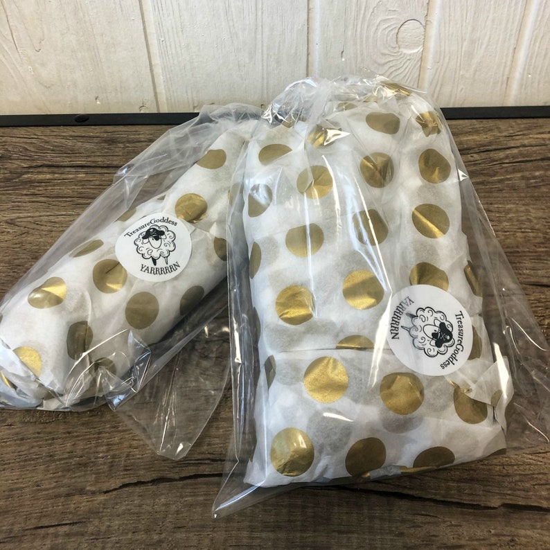 May include: Two wrapped packages in clear plastic bags. The packages are wrapped in white paper with gold polka dots. Each package has a round sticker with the text "Treasure Goddess" and "YARRRRRN".