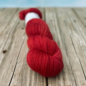 May include: A skein of red yarn with a white section at the top, resting on a weathered wooden surface. The yarn is tightly wound, showing a textured surface. The background is a soft blue, creating a contrast with the yarn's color.
