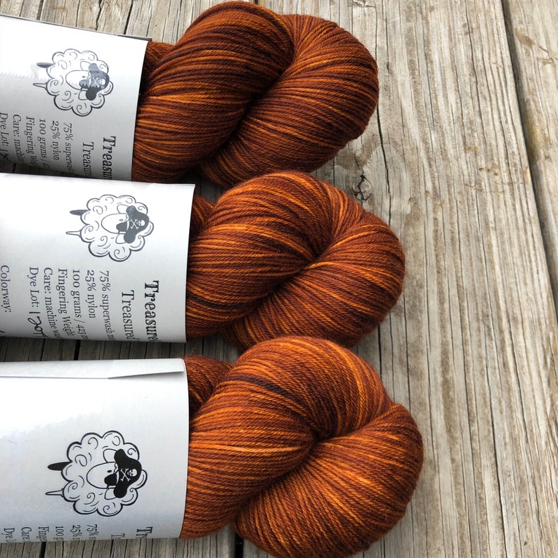 May include: Three skeins of brown and orange Treasure yarn, each with a label featuring a sheep with a skull and crossbones. The yarn is 75% superwash and 25% nylon, 100 grams/423 yards. The colorway is 1201.