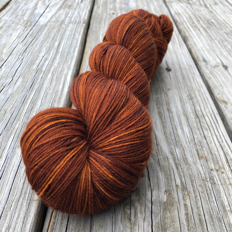 May include: A skein of brown yarn with orange highlights, resting on a weathered wooden surface. The yarn is tightly wound, showcasing a gradient of warm, earthy tones. Ideal for knitting or crochet projects.