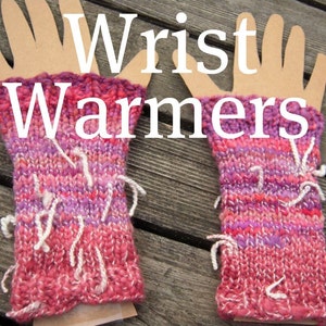 May include: A pair of hand-knitted wrist warmers in shades of pink, red, and purple. The text "Wrist Warmers" is displayed in white across the top. The warmers have white fringe details and are displayed on a wooden surface.