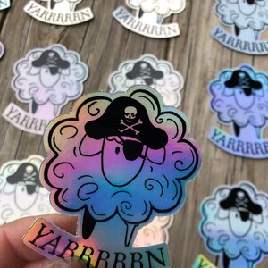 May include: Holographic sticker featuring a cartoon sheep wearing a pirate hat with a skull and crossbones. The sheep has a fluffy, cloud-like body and the word "YARRRRRN" is printed below. The sticker has a rainbow sheen.