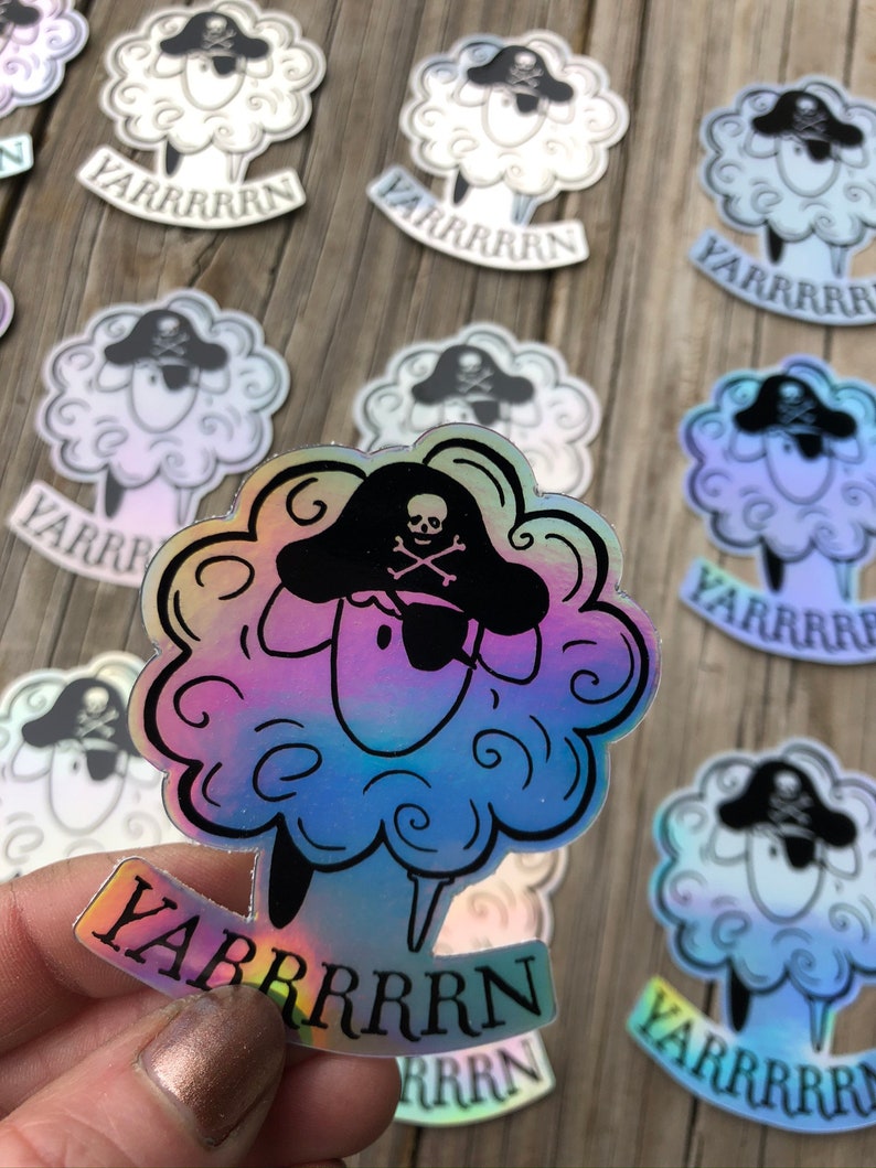 May include: Holographic sticker featuring a cartoon sheep wearing a pirate hat with a skull and crossbones. The sheep has a fluffy, cloud-like body and the word "YARRRRRN" is printed below. The sticker has a rainbow sheen.