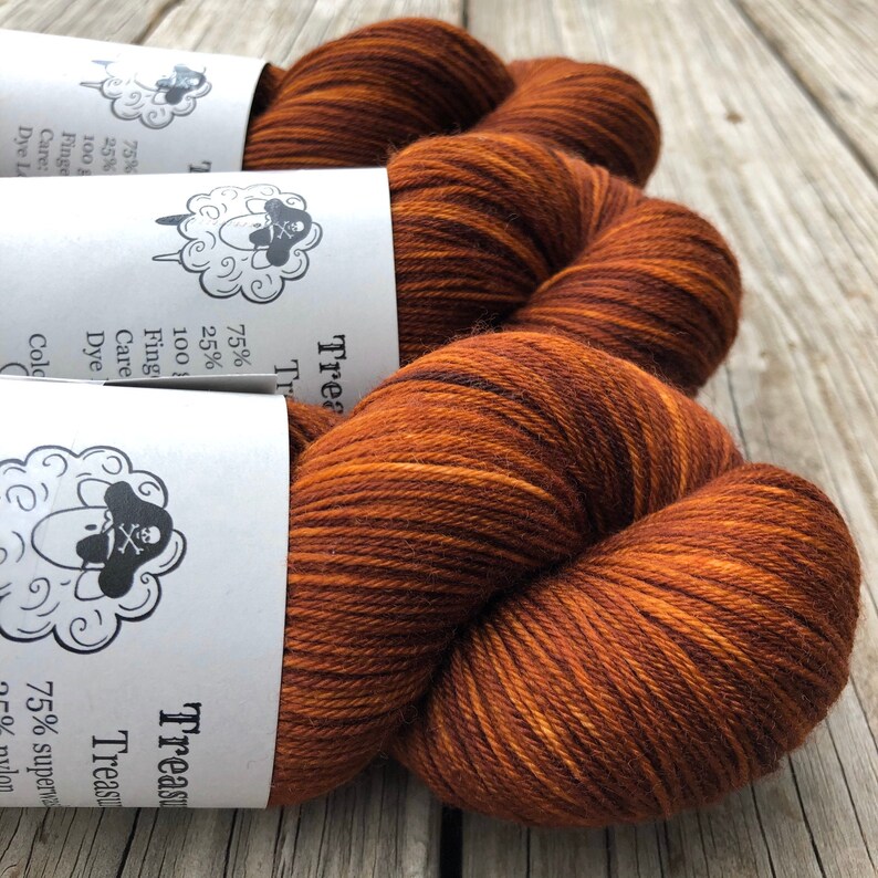 May include: Close-up of three skeins of Treasure yarn in shades of brown and orange. The yarn is tightly wound, with visible texture and color variations. Each skein has a white label with a sheep logo and text.