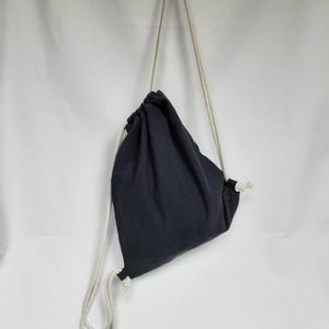 Könnte beinhalten: Ein marineblauer Rucksack mit Kordelzug und cremefarbenen Seilriemen. Die Tasche ist aus einem weichen Stoff gefertigt und hängt vor einem weißen Hintergrund. Die Tasche ist oben mit dem Kordelzug gerafft.