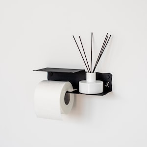 Könnte beinhalten: Schwarzer Toilettenpapierhalter aus Metall mit einem Regal und einem weißen Keramikdiffusor mit schwarzen Schilfrohren. Der Halter ist an einer weißen Wand montiert.