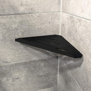 May include: A black, triangular corner shelf made of metal. The shelf is mounted on a grey tiled wall and features a slotted design. The shelf is designed to fit into a corner and provide storage space.