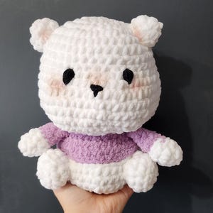 Può includere: Un orsetto bianco all'uncinetto fatto a mano con un maglione lavanda. L'orso ha occhi neri, un naso nero e guance rosa. È un giocattolo morbido e di peluche.