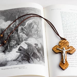 May include: A wooden cross pendant with a silver figure of Jesus Christ hangs on a brown cord. The cross is resting on an open book with text and a black and white illustration of angels. The pendant is a religious item.