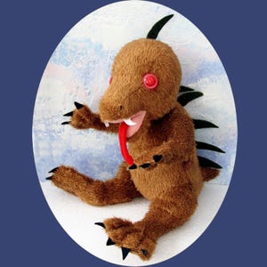 May include: A brown plush toy creature with red button eyes, a long red tongue, and black claws. The handmade monster has green spikes down its back and is set against a blue and white background.