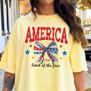 May include: A light yellow t-shirt with the word "AMERICA" in red, a bow in red, white, and blue, and the text "1776 land of the free." The shirt is a casual style.