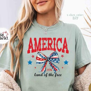 May include: A light green t-shirt with the word "AMERICA" in red block letters. Below the word is a bow in red, white, and blue, with the text "1776 land of the free". The shirt has a relaxed fit.