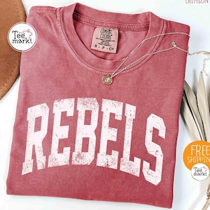 Puede incluir: Camiseta de color carmesí con la palabra "REBELS" en letras blancas desgastadas. La camiseta tiene cuello redondo y una pequeña etiqueta. Un collar dorado con un colgante está drapeado sobre la camiseta. Un sombrero marrón y una planta seca están en el fondo.