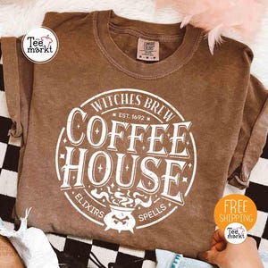 Könnte beinhalten: Ein braunes T-Shirt mit einem weißen Aufdruck, der "Witches Brew Coffee House" mit einer Kessel-Illustration zeigt. Das Shirt hat auch den Text "Elixirs" und "Spells". Das Shirt ist von der Marke Comfort Colors.