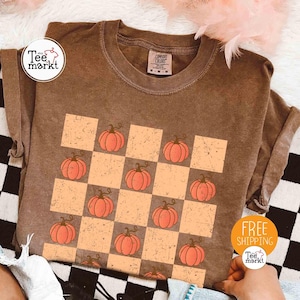 Könnte beinhalten: Braunes T-Shirt mit einem Schachbrettmuster aus orangefarbenen Quadraten und Kürbissen. Das Shirt hat einen Rundhalsausschnitt und kurze Ärmel. Der Text "Tee Markt" befindet sich auf einem weißen Kreis.