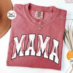 May include: Crimson t-shirt featuring the word "MAMA" in a baseball-style font. The letters are white with red stitching. A gold necklace and a decorative plant are also visible.
