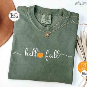Peut inclure: T-shirt vert mousse avec l'inscription "hello fall" en écriture blanche et un motif de citrouille orange. Un collier doré avec un pendentif est posé sur le t-shirt. Le t-shirt est plié sur une surface blanche.