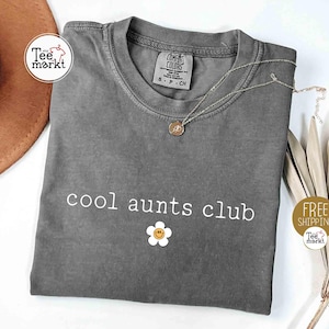 Peut inclure: Un t-shirt gris avec l'inscription blanche "cool aunts club" et un motif de marguerite souriante. Un collier argenté avec un pendentif est posé sur le t-shirt. La marque du t-shirt est Comfort Colors.
