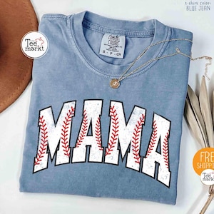 May include: A blue jean coloured t-shirt with the word "MAMA" in white block letters with red baseball stitching. A gold necklace with a pendant rests on the shirt. The shirt has a crew neck and short sleeves. The shirt is from Comfort Colors.