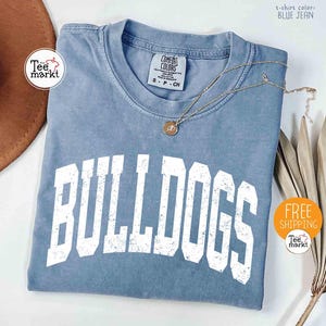 May include: A blue jean colored t-shirt with the word "BULLDOGS" in large white block letters. A gold necklace with a pendant rests on the shirt. The shirt has a Comfort Colors tag. A brown hat and dried plant are in the background.