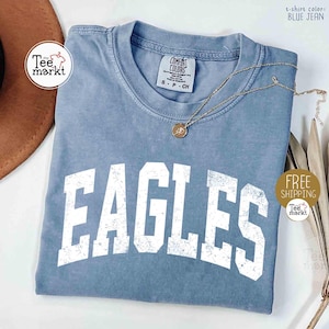 May include: A blue jean colored t-shirt with the word "EAGLES" in large white block letters. The shirt has a crew neck and short sleeves. A gold necklace with a pendant is draped across the shirt. The text "t-shirt color: BLUE JEAN" is visible.