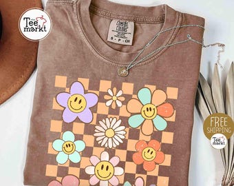 May include: Brown t-shirt with a retro floral design featuring smiley faces and a checkered background. The shirt has a crew neck and short sleeves. A silver necklace with a pendant is draped across the shirt. The shirt has the text "Comfort Colors" on the tag.