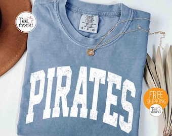 May include: A blue jean-colored t-shirt with the word "PIRATES" in large white block letters. The shirt has a crew neck and short sleeves. A gold necklace with a pendant rests on the shirt. The shirt is from Comfort Colors.