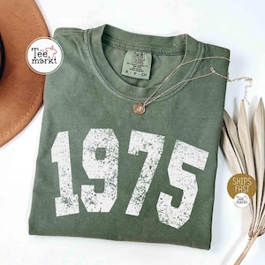 May include: A green t-shirt with the year 1975 printed in white distressed font. The shirt is folded and laid flat on a white surface. The shirt has a tag that says 'Comfort Colors' and 'S - P - CH'.
