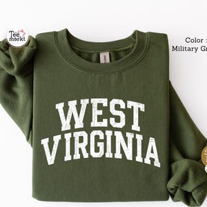 May include: A military green long-sleeve crewneck sweatshirt featuring the text WEST VIRGINIA printed in a distressed white font across the chest. The garment has a ribbed collar, cuffs, and hem, with a standard fit and a soft fleece interior.