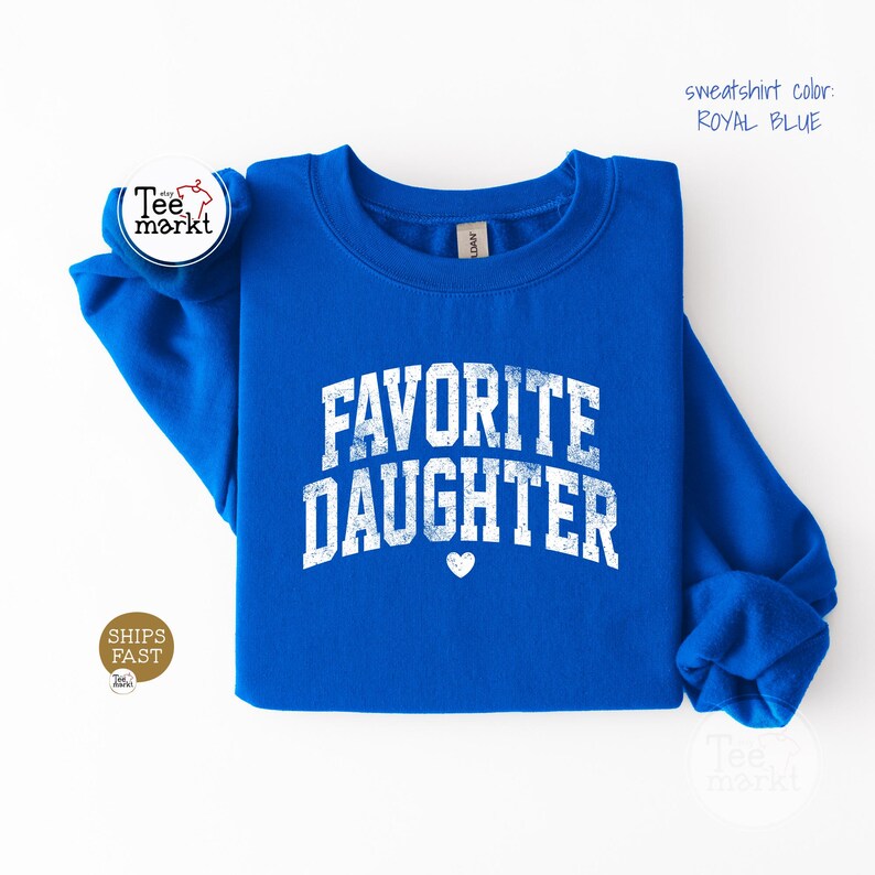 May include: A royal blue long-sleeve sweatshirt featuring the white distressed text "FAVORITE DAUGHTER" in an arched collegiate font with a small white heart icon centred below the text. The garment has a crew neckline and long sleeves.