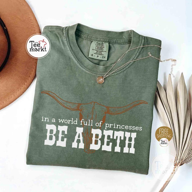 Puede incluir: Camiseta verde oliva con el texto blanco "in a world full of princesses BE A BETH" y un gr&aacute;fico de calavera de cuernos largos marr&oacute;n. Tambi&eacute;n se ven un collar dorado y un sombrero marr&oacute;n.