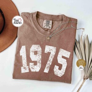 May include: A brown t-shirt with the year "1975" printed in large, white, distressed lettering. A silver necklace with a pendant rests on the shirt. The shirt is from the "Comfort Colors" brand and has a small tag. A brown hat is visible in the background.